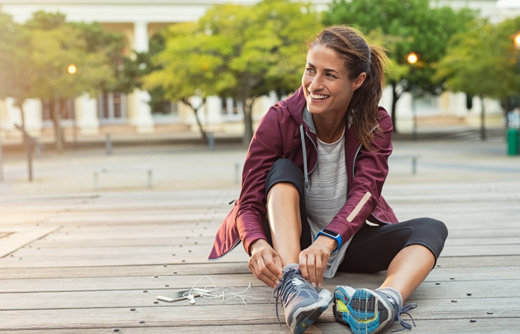 Mature fitness woman tie shoelaces on road. Cheerful runner sitting on floor on city streets with mobile and earphones wearing sport shoes. Active latin woman tying shoe lace before running.