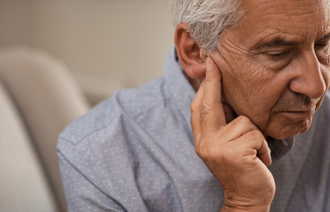 Side view of senior man with symptom of hearing loss. Mature man sitting on couch with fingers near ear suffering pain.