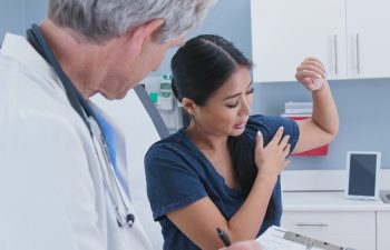 Japanese woman explaining shoulder pain to doctor in exam room. Patient with rotator cuff injury talking to medical professional