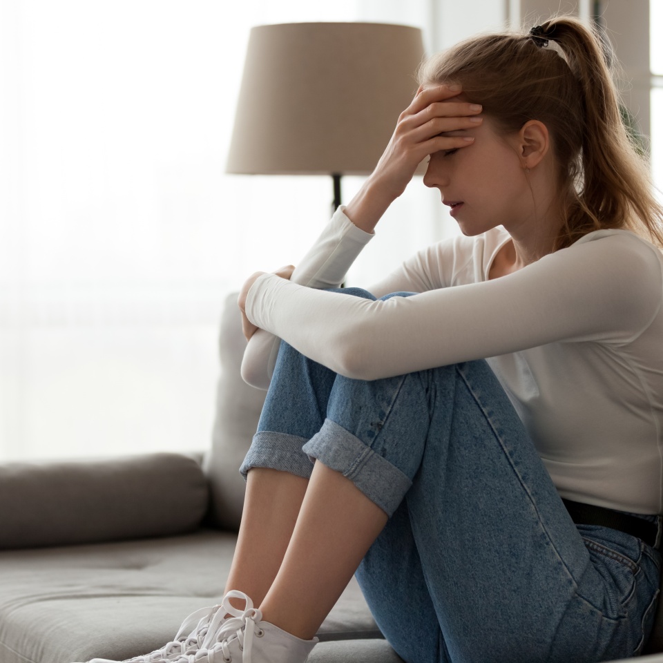 Upset woman frustrated by problem with work or relationships, sitting on couch, embracing knees, covered face in hand, feeling despair and anxiety, loneliness, having psychological trouble