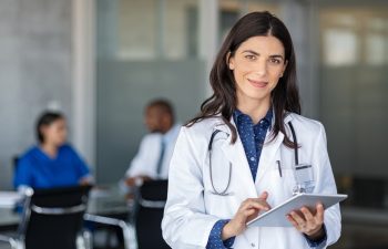 Portrait of beautiful mature woman doctor holding digital tablet and looking at camera. Confident female doctor using digital tablet with colleague talking in background at hospital. Latin nurse.