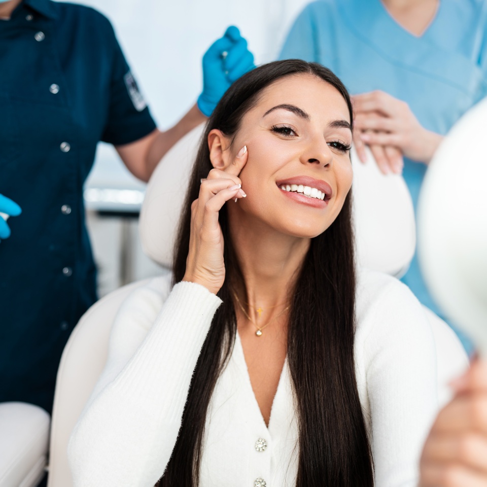 Beautiful and happy young woman sitting in medical chair and looking in the mirror. She is satisfied after successful beauty treatment with hyaluronic acid fillers or botulinum toxin injections.