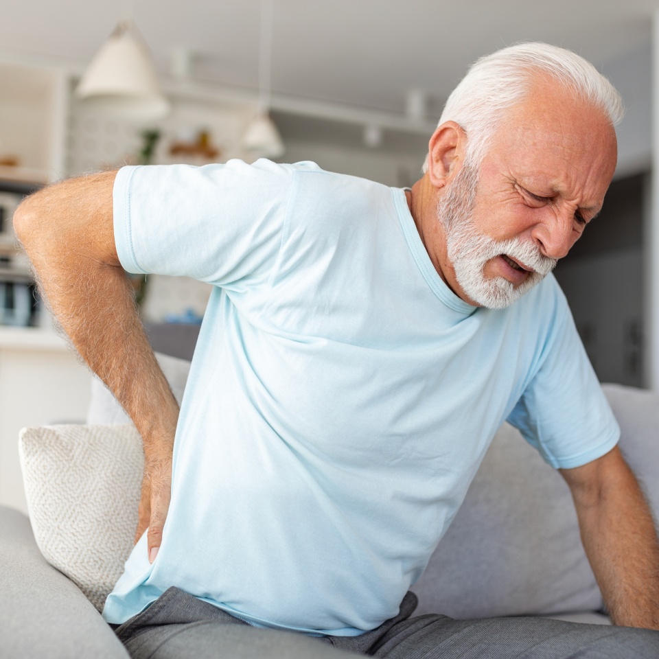 Senior man, with back pain, touches his back, illustrating sciatica and sedentary lifestyle. Emphasizing spine health and the significance of healthcare and insurance in this stock image.