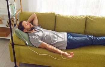 Side view of smiling man lying with closed eyes on the couch at home or in medical clinic while receiving IV drip infusion and vitamin therapy in his blood. Person receiving injection therapy.