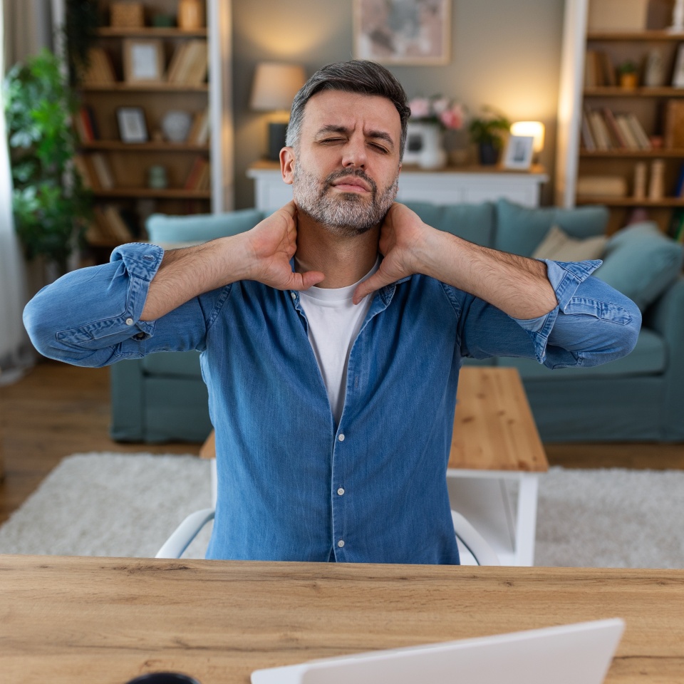 Businessman feeling pain in neck after sitting at the table with laptop. Tired man suffering of office syndrome because of long hours computer work. He is massaging his tense neck muscles
