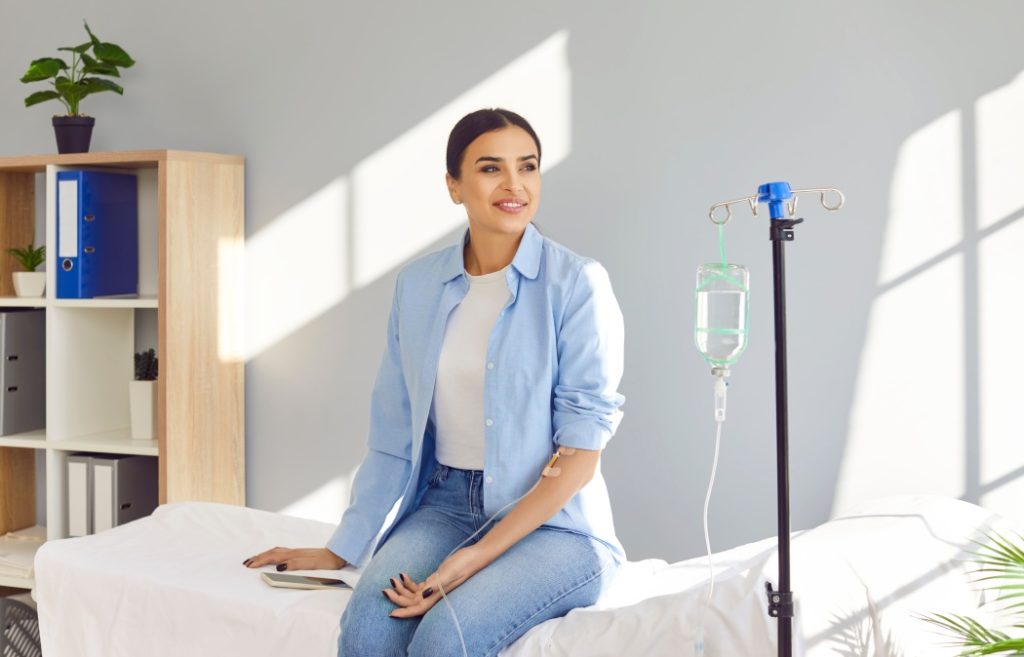 Young happy smiling woman sitting on the couch in medical clinic looking at the window while receiving IV drip infusion and vitamin injection therapy in her blood. Healthcare concept.