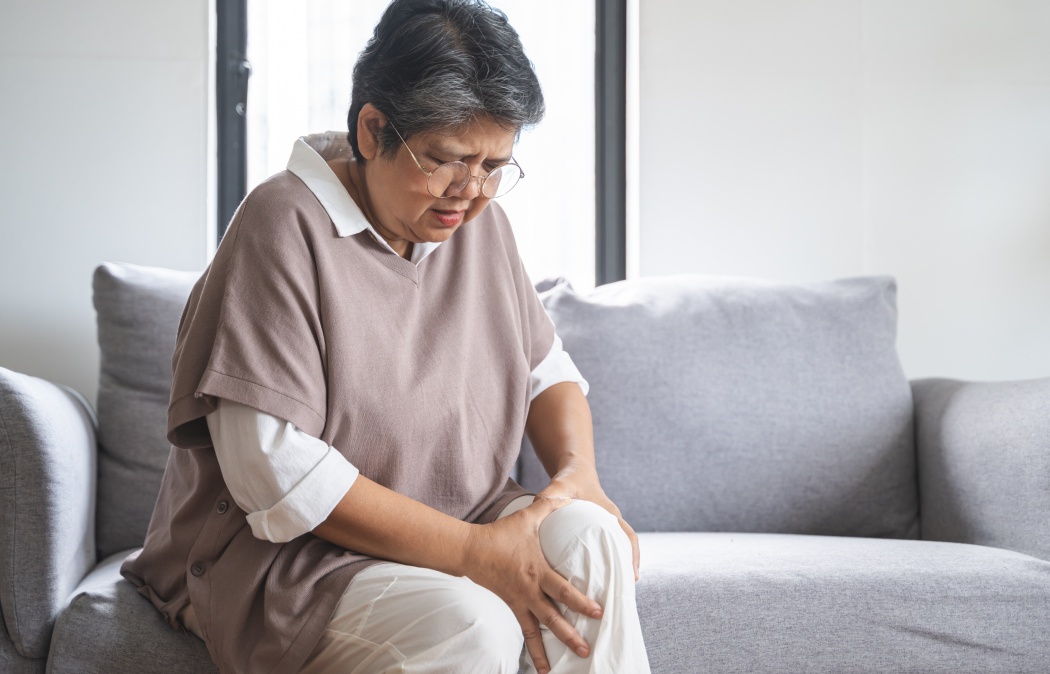 Body arthritis muscles stiff problem, aged woman suffering on leg, pain knee from disease chronic, hand massage rubbing joint injury, mature hurt or sore sitting on sofa at home. Health care people.