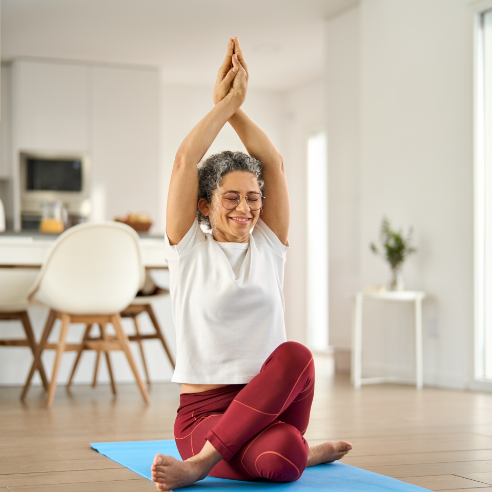 Happy healthy fit mature woman practicing yoga doing exercises sitting on mat at home interior. Sporty mindful older middle aged lady in her 50s stretching body with eyes closed in house living room.