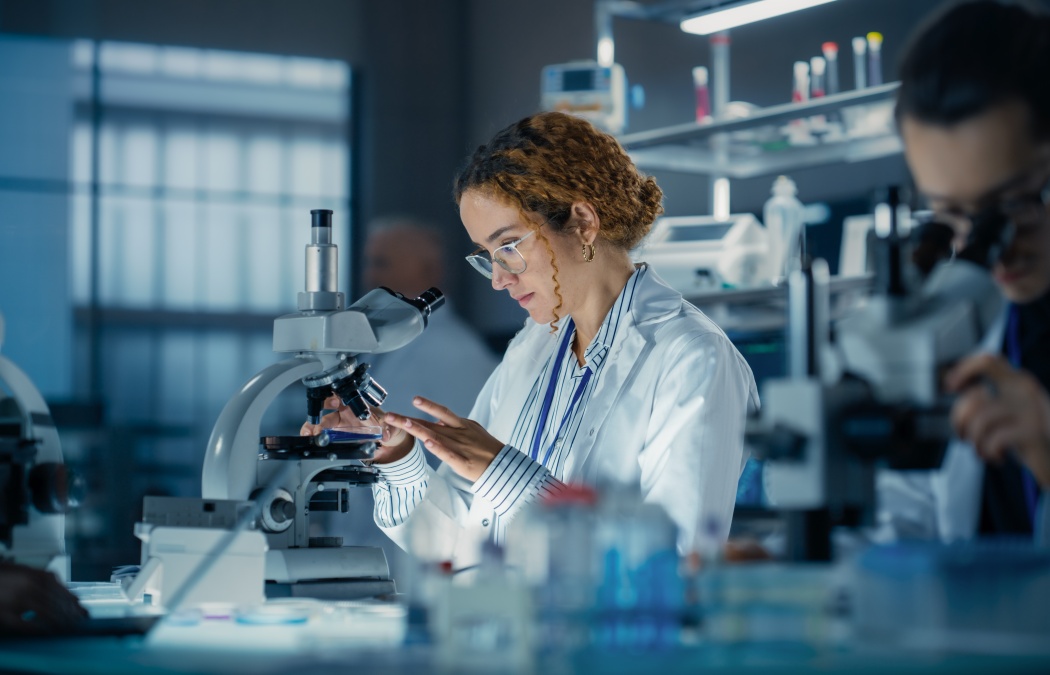 Hisplanic Female Bioengineer Looking at DNA Samples Under a Microscope in a Modern Applied Science Laboratory. Portrait of a Young Lab Engineer in White Coat Inventing New Medical Drugs