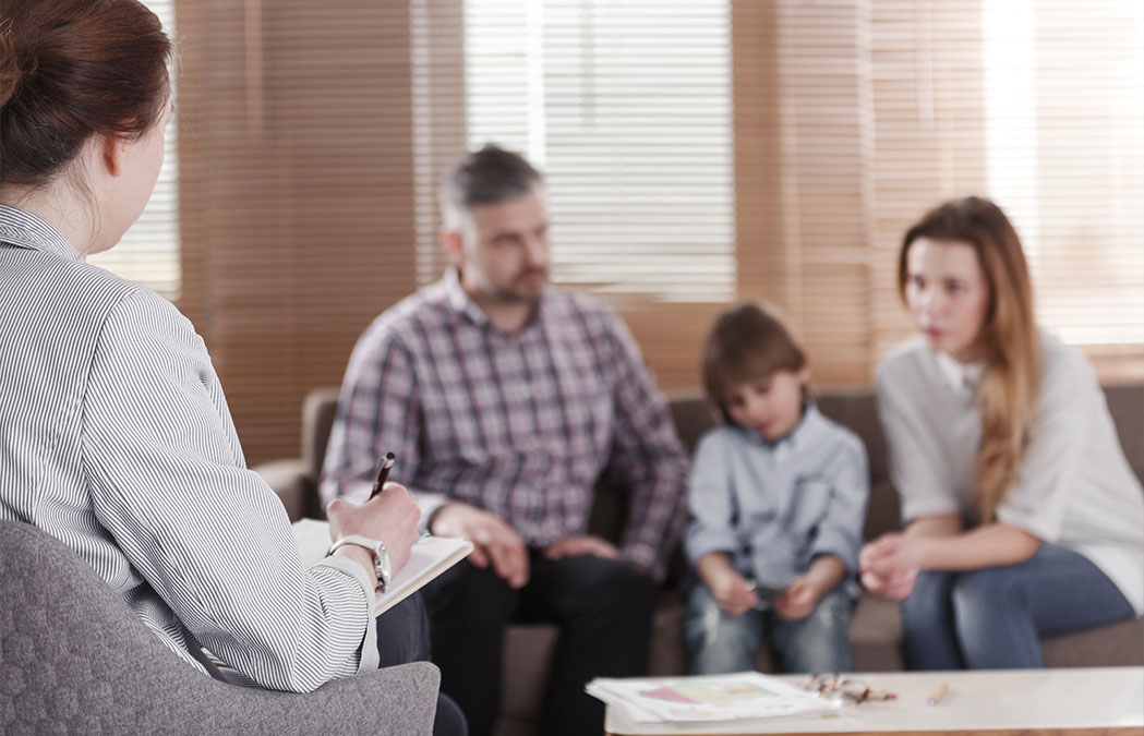 A therapist takes notes while talking with a family of three, including two adults and a child, seated together on a couch in an office.