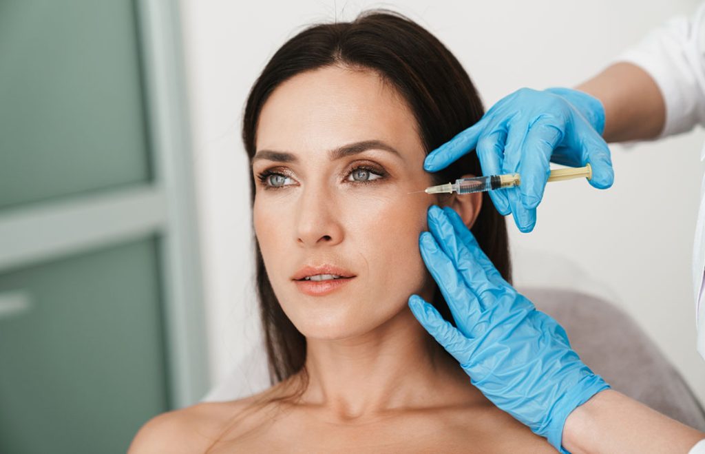 A woman receives a cosmetic facial injection near her cheek from a medical professional wearing blue gloves.