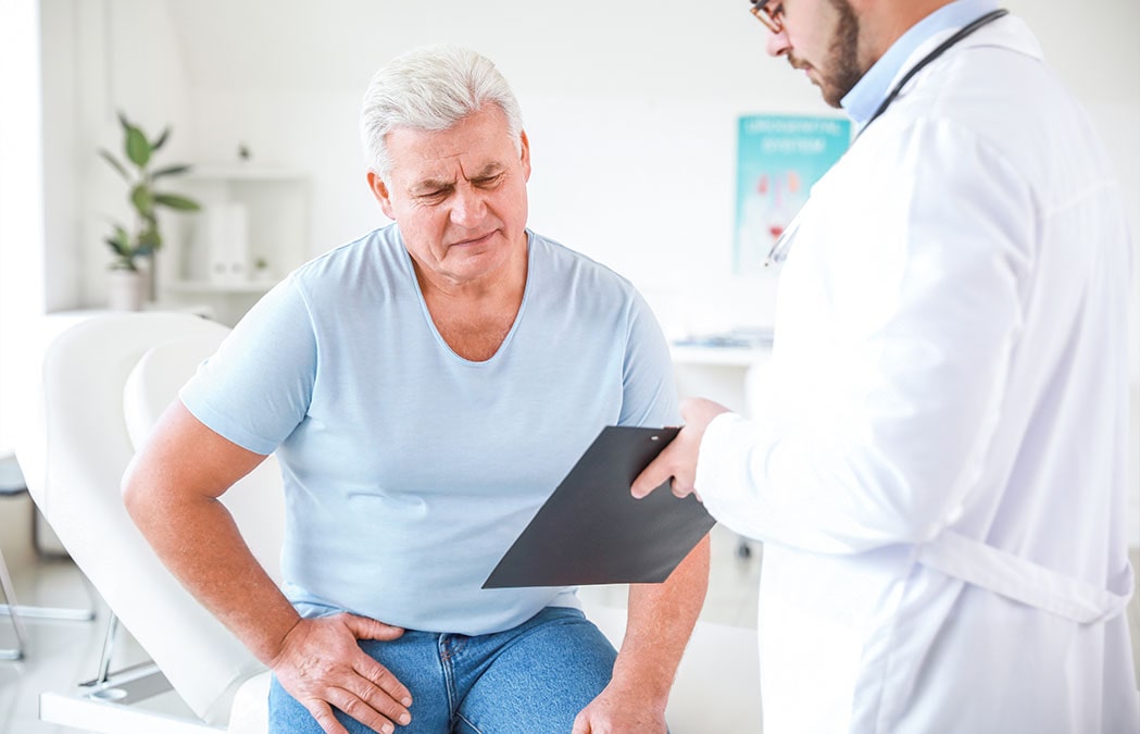 An older man sits on an exam table holding his hip while a doctor in a white coat shows him a clipboard, suggesting a medical consultation.