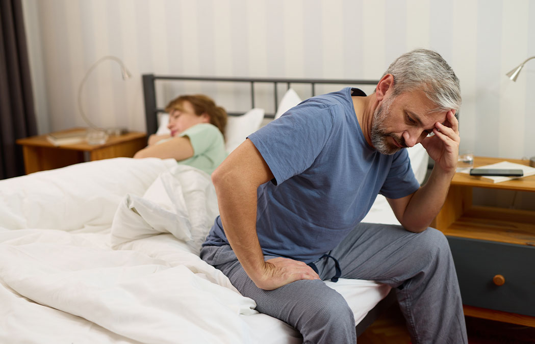 A middle-aged man sits on the edge of a bed holding his head, looking stressed, while a woman sleeps behind him under the covers.