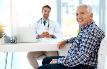 An older man in a plaid shirt sits at a desk, smiling, while a doctor in a white coat sits across from him with a laptop and paperwork.