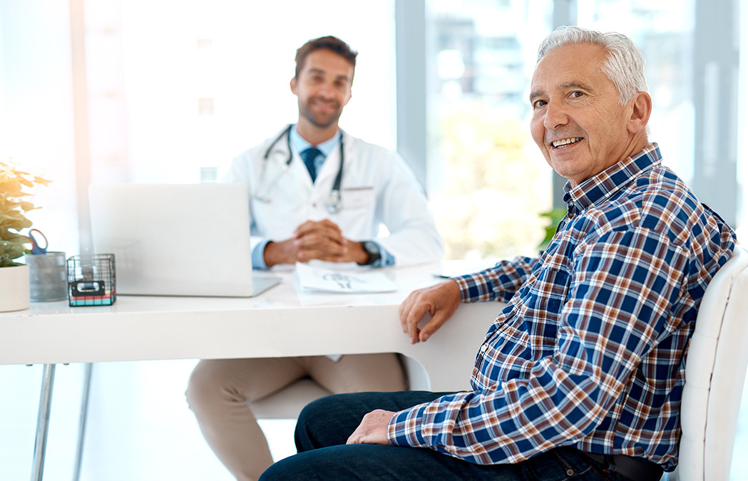An older man in a plaid shirt sits at a desk, smiling, while a doctor in a white coat sits across from him with a laptop and paperwork.