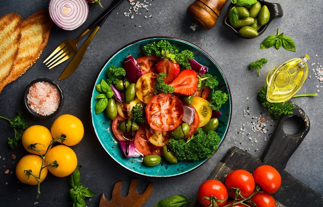 A colorful salad with tomatoes, strawberries, red onion, olives, parsley, and broccoli in a blue bowl, surrounded by bread, salt, olive oil, and fresh vegetables on a dark surface.