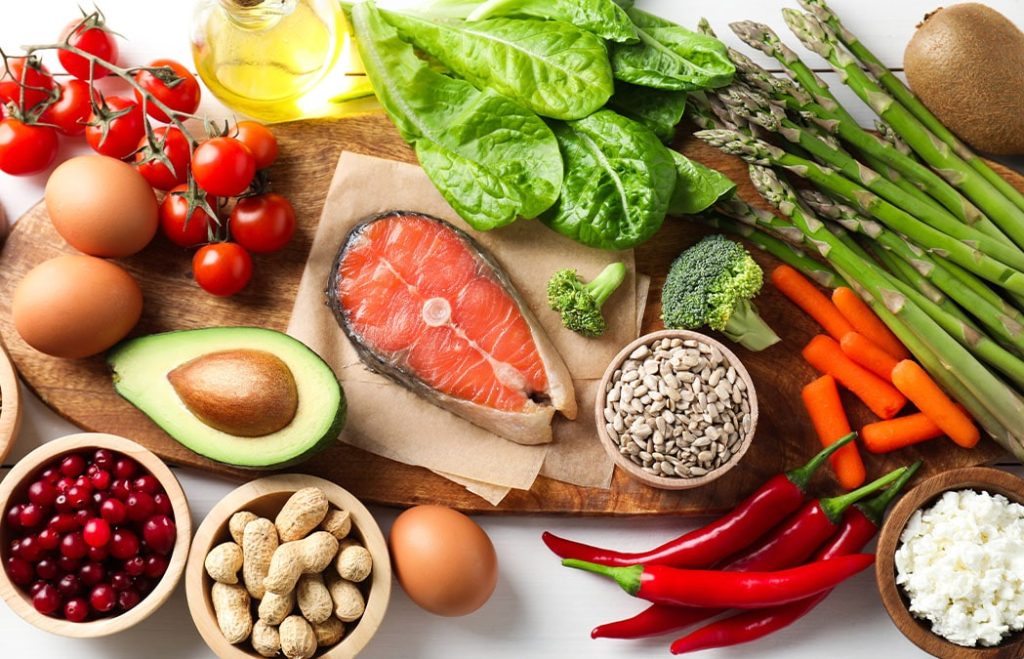 A variety of fresh foods including salmon, avocado, eggs, tomatoes, spinach, asparagus, carrots, broccoli, sunflower seeds, peanuts, cranberries, and red chili peppers on a wooden board.