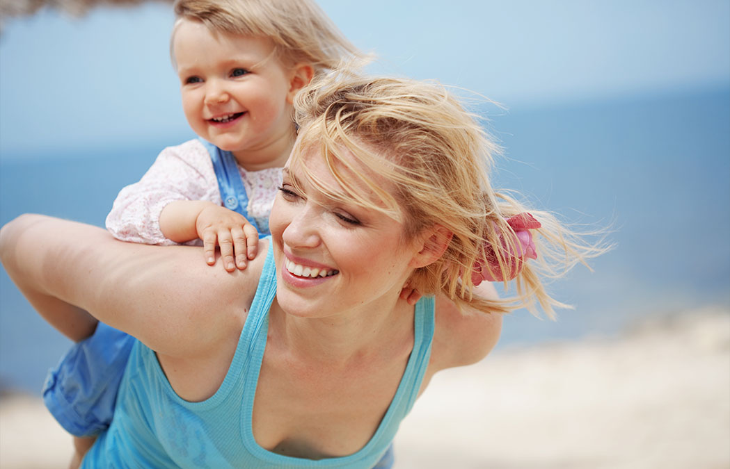 A woman gives a piggyback ride to a smiling toddler on a beach, with the ocean and sand visible in the background.