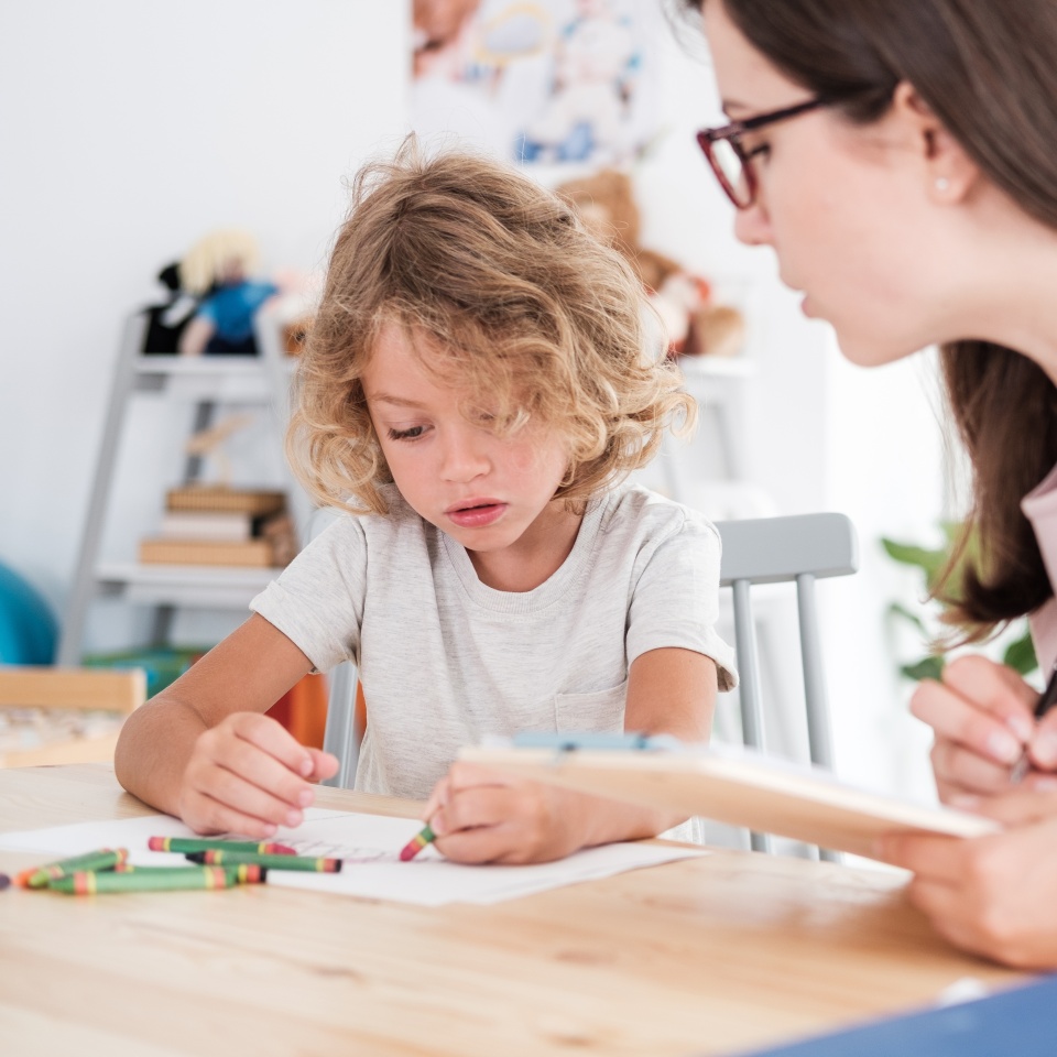 A psychotherapist watching a child draw pictures with crayons during an evaluation in a private school.