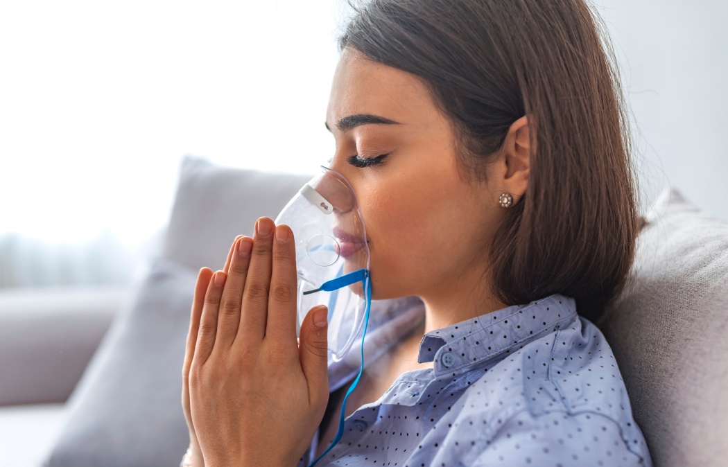 Use nebulizer and inhaler for the treatment. Young woman inhaling through inhaler mask lying on the couch. Woman using asthma machine at home
