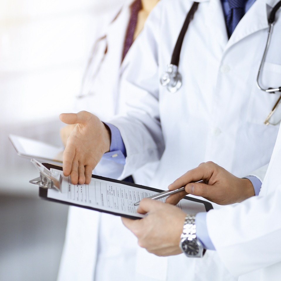 Unknown doctors, two men and a woman, discuss medical exam resoults, while standing at sunny hospital office. Physicians using clipboards for filling up medication history records. Perfect medical