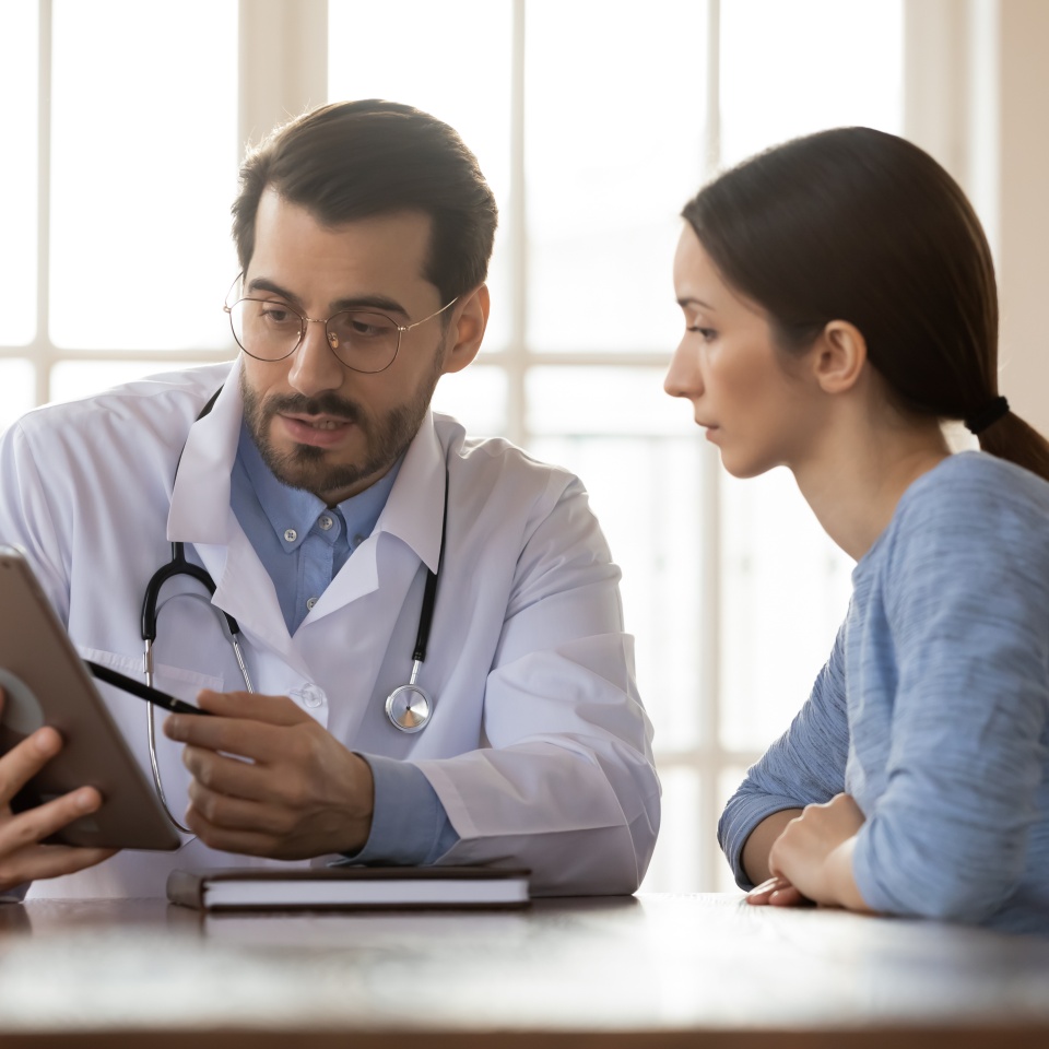 Focused young male doctor pointing at digital computer tablet screen, sharing health tests laboratory results to serious female patient or explaining medical insurance benefits at meeting in clinic.