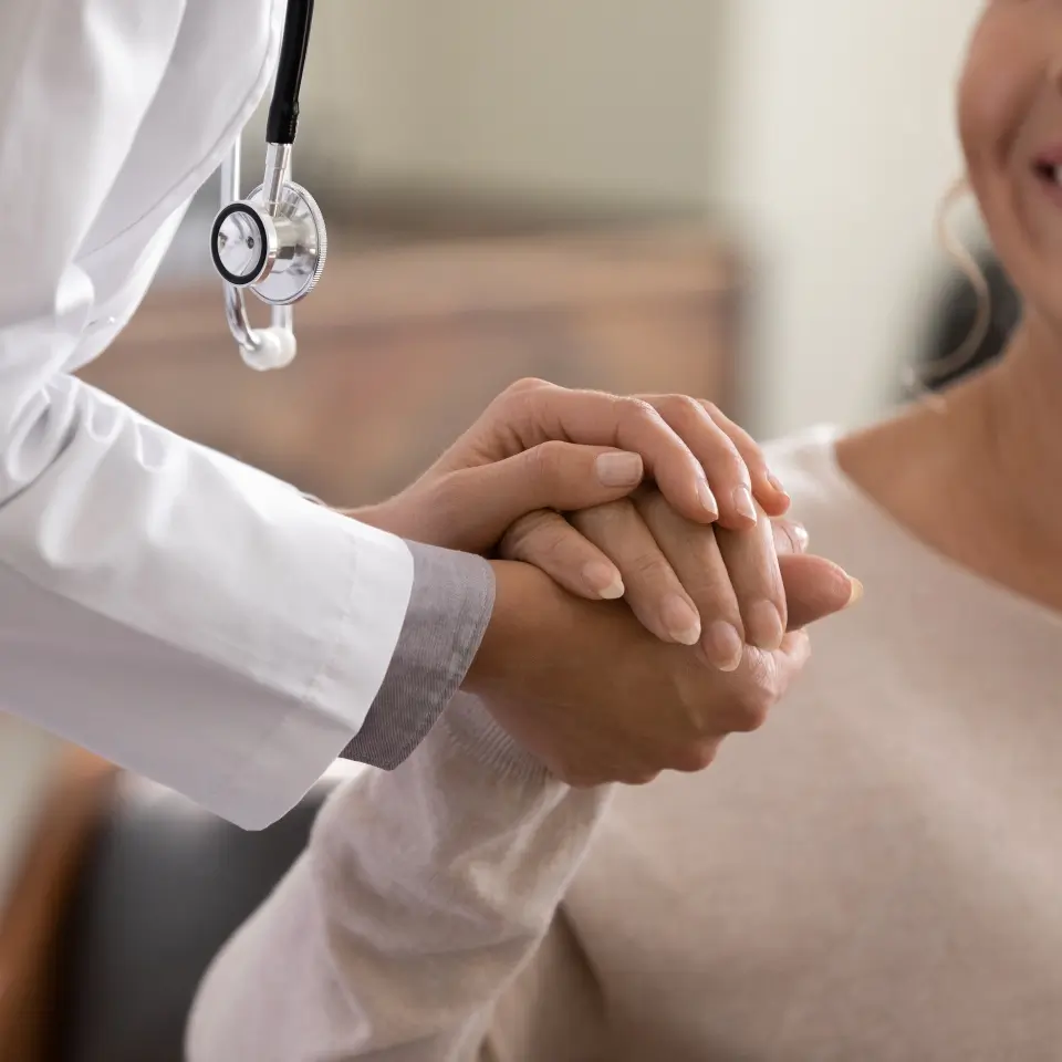 A doctor wearing a white coat holds a patient's hand in a reassuring gesture; only their hands and part of their upper bodies are visible.