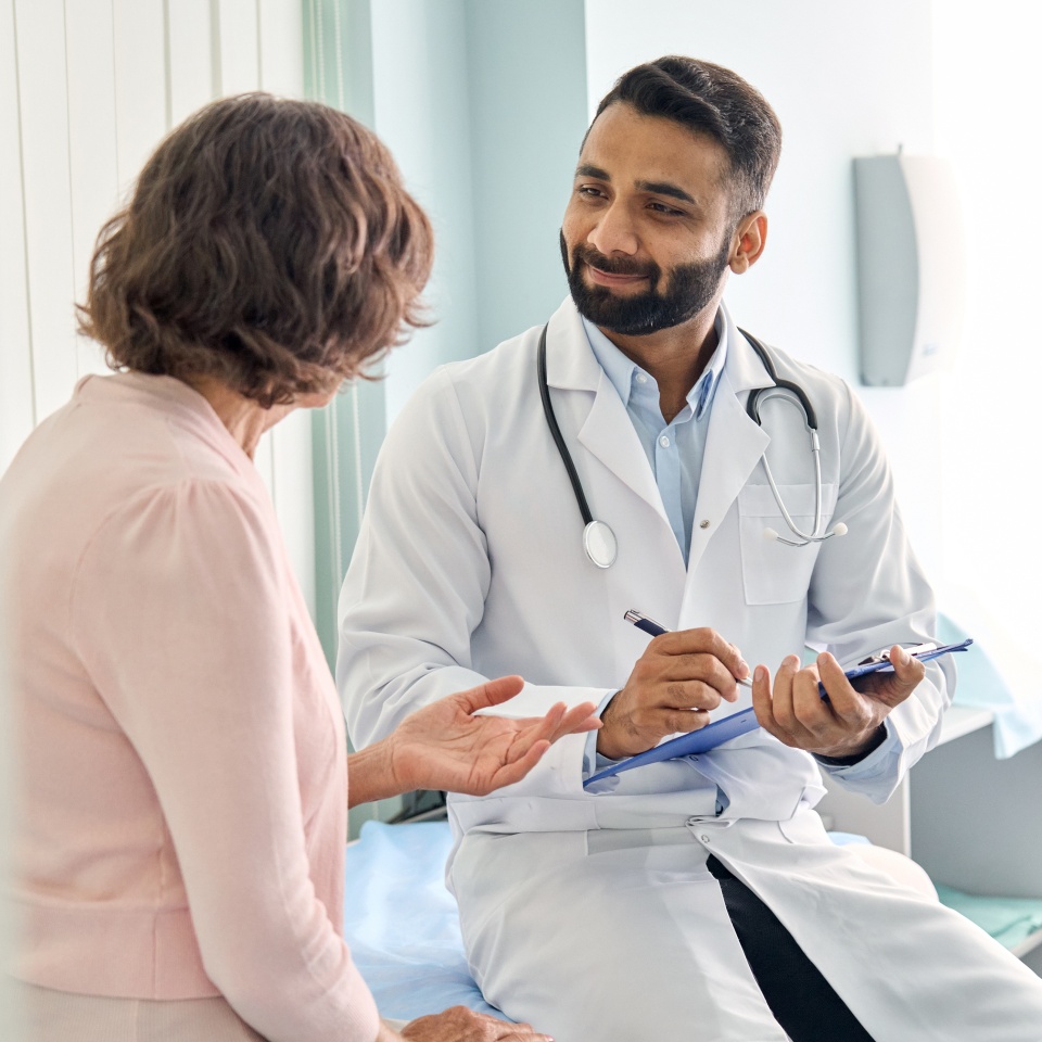 Indian male doctor consulting senior old patient filling form at consultation. Professional physician wearing white coat talking to mature woman signing medical paper at appointment visit in clinic.