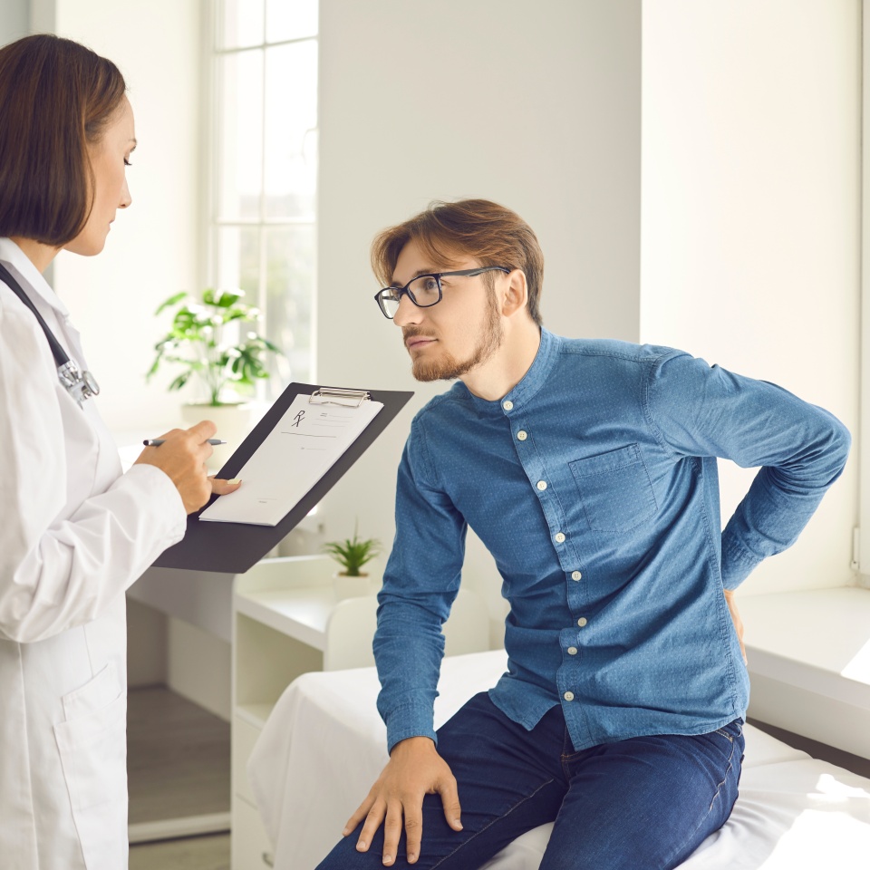 Young man tells doctor about backache while sitting on examination couch during visit to clinic. Male patient listening to physician giving advice and explaining how to treat bad pain in injured back
