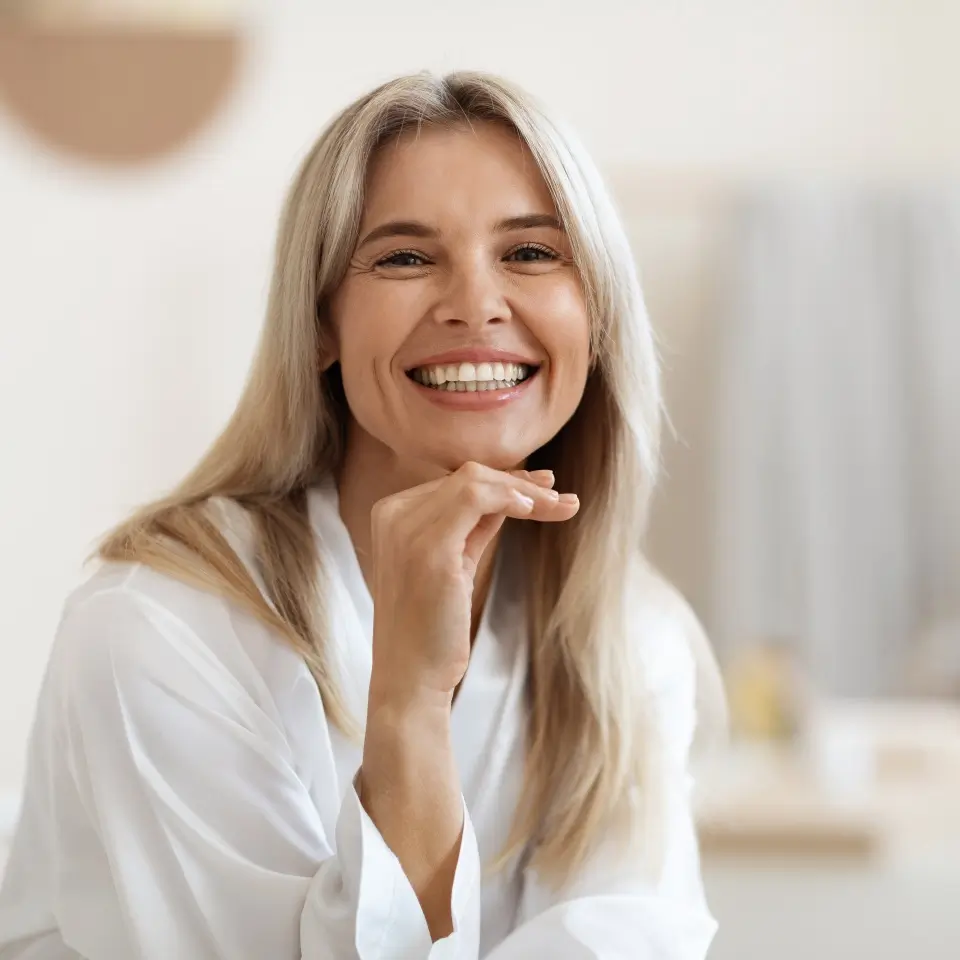 A woman with long blonde hair wearing a white robe smiles at the camera with her hand resting under her chin.