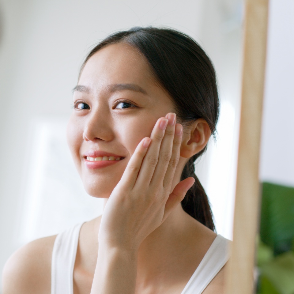 Young Asian woman touching healthy facial beauty skin look at mirror