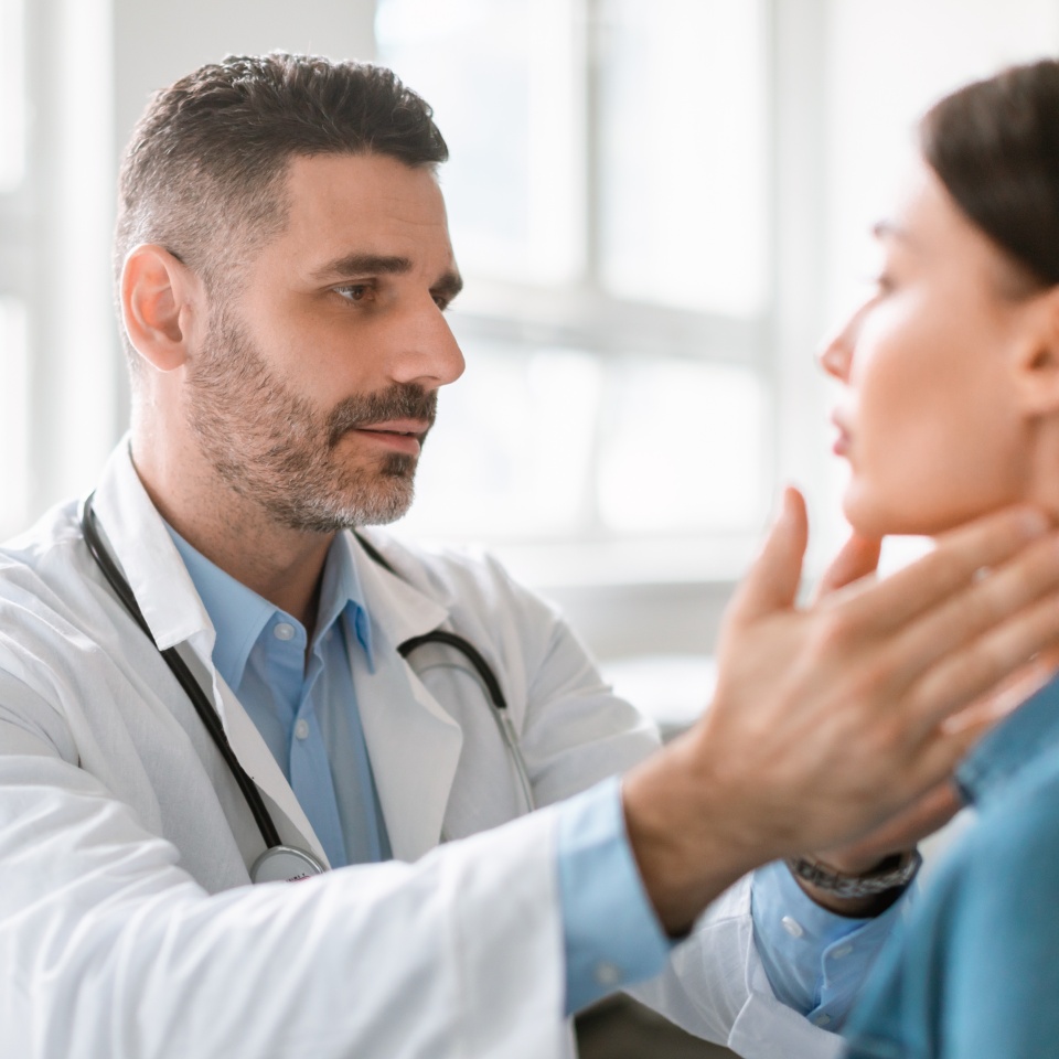 Friendly middle aged male doctor checking sore throat or thyroid glands, touching neck of young female patient visiting clinic office. Thyroid cancer prevention concept