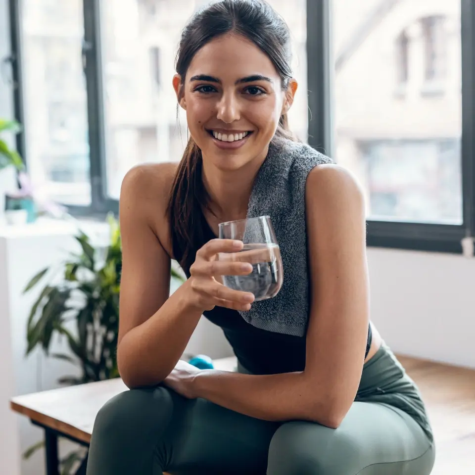 A woman in workout clothes sits indoors with a towel around her neck, smiling and holding a glass of water.