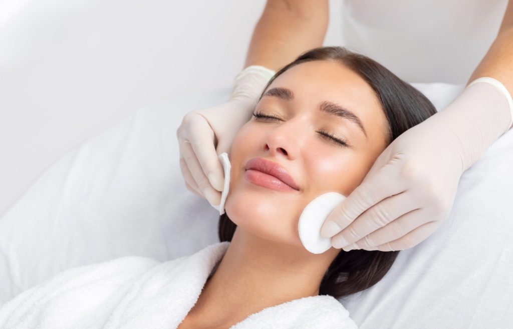 Female beautician cleansing skin of young woman, cosmetology procedure in beauty salon. Anti-age procedures at spa clinic