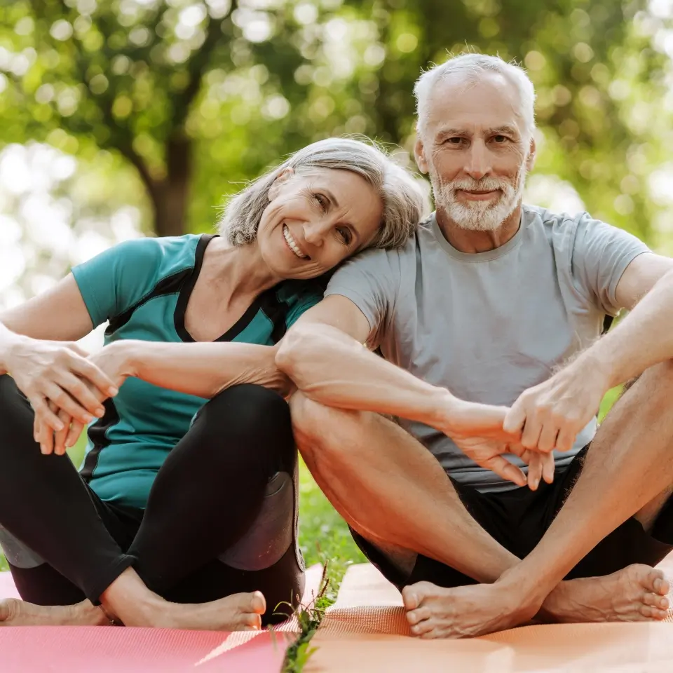 An older couple sits on yoga mats outdoors, the woman leaning her head on the man's shoulder, both smiling, surrounded by greenery.
