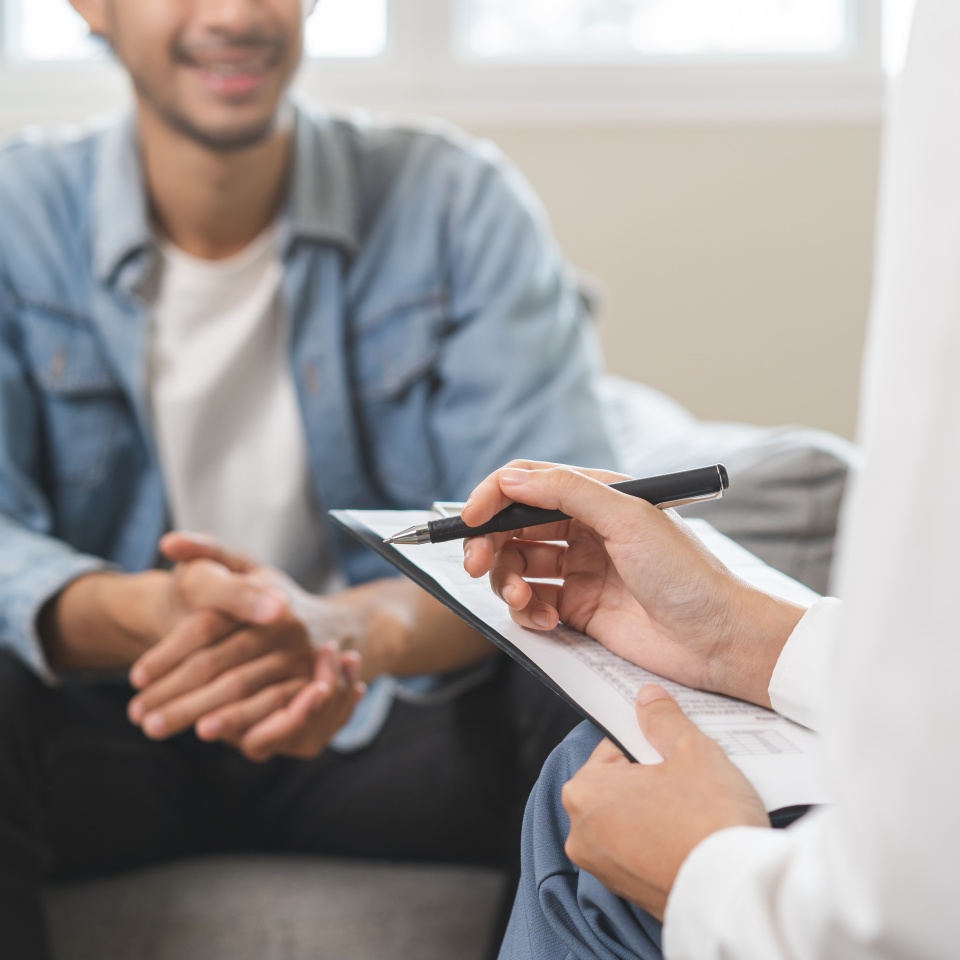 Psychology, depression. Happy smile asian young man patient mental therapy explain symptom with psychologist while doctor woman taking notes at clinic. Psychologist with sick mental health person.