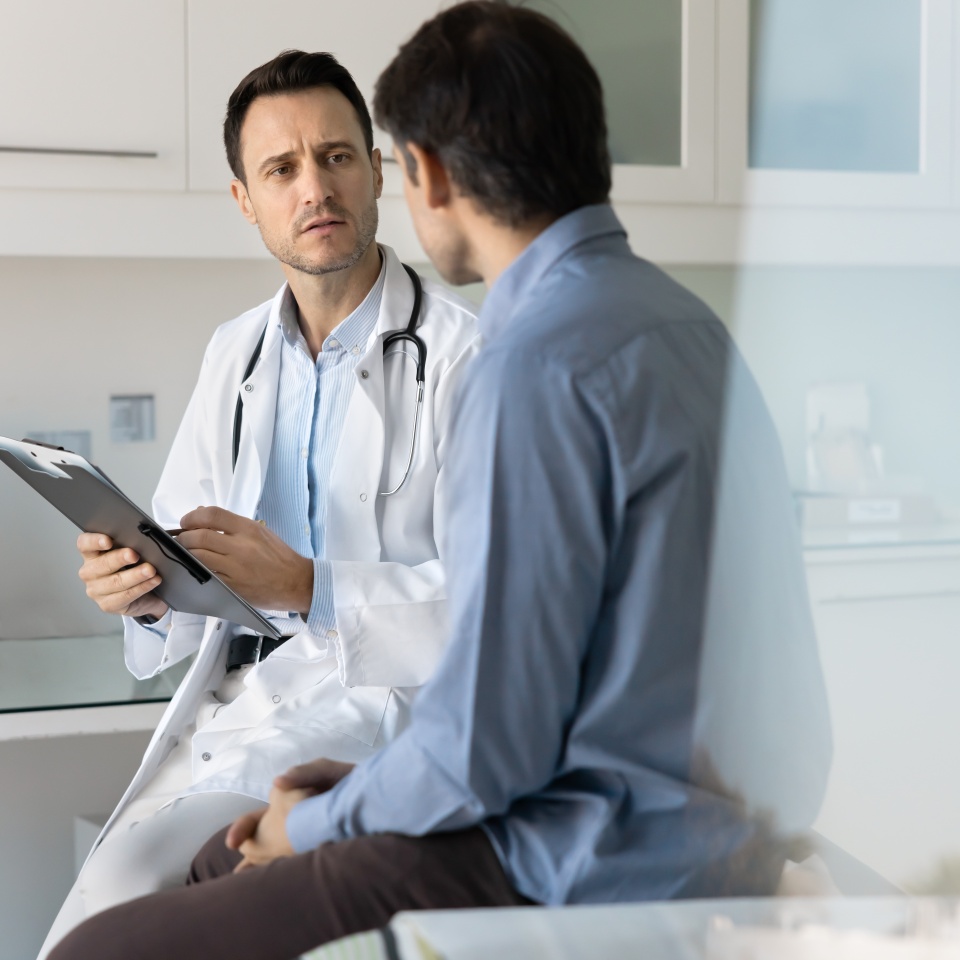 Focused concerned doctor examining male patient, working in clinic office, writing notes, records on pad, talking to man about healthcare complaints, sitting on couch. Vertical candid shot