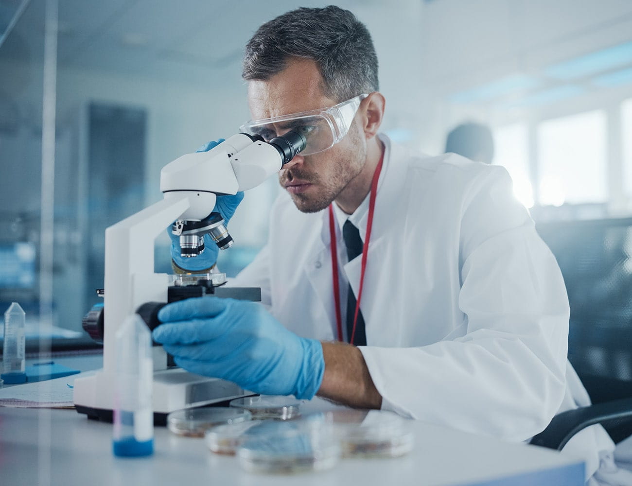 A scientist wearing safety goggles and gloves examines samples under a microscope in a laboratory setting.
