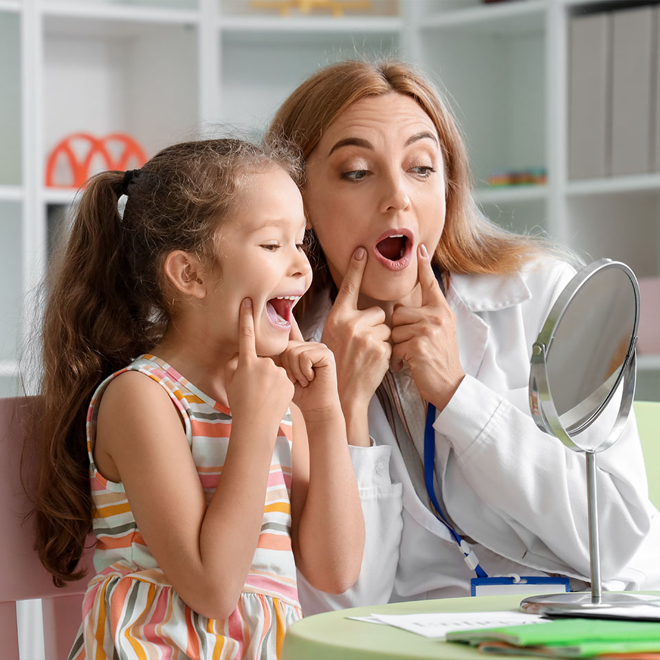 Mature speech therapist working with little girl near mirror at table in office