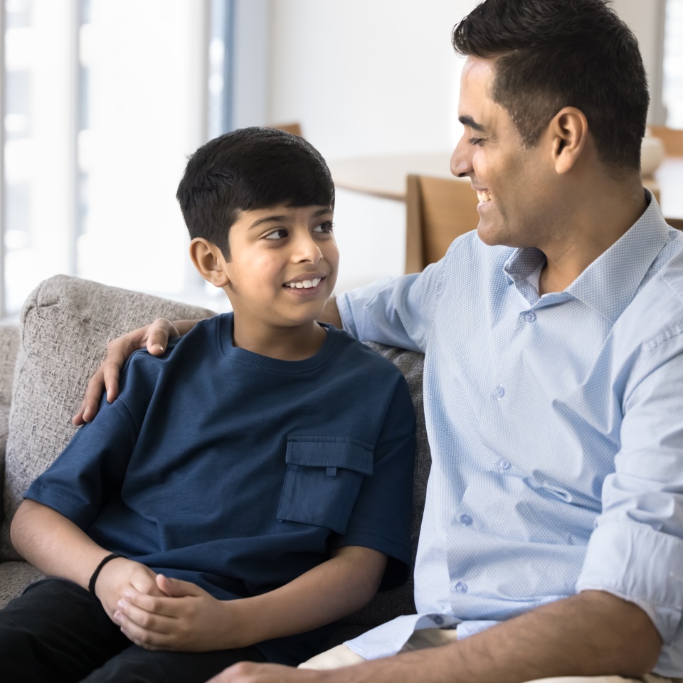 Happy Indian preteen son talking to loving father on home sofa. Positive caring dad enjoying friendship, good relationship with teenage kid, giving support to boy, hugging child